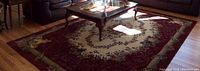 Full view of the burgundy Cameo Rose area rug placed on a hardwood floor under a glass coffee table, showing the detailed floral design and border pattern.