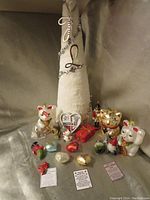 Photo showing a white fabric cone-shaped Christmas tree decorated with silver garland and a silver 'L' initial at top. Surrounding the tree are four lucky cat figurines in white, gold, and red, two heart-shaped ornaments, a nutcracker figurine, two small gnomes, and red gift box decor, all placed on a gray backdrop.
