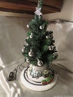 Photo of the tree showing the full Christmas tree decorated with silver ball ornaments and star topper, set on a wintery village base with a toy train on a circular track.