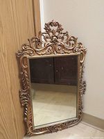 Full view of vintage gold ornate wall mirror leaning against wall, showing detailed decorative frame and reflective mirror surface.