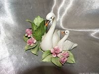 Porcelain figurine of two white swans with surrounding pink roses and green leaves, positioned on a silver satin fabric background