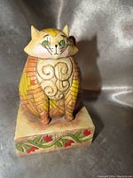 Front and side view of the stylized fat cat figurine showing the detailed face, front pattern and decorative base with floral motifs.