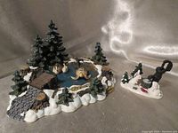 Wide view of Pine Haven Pond scene with two swans on pond, pine trees, bridge, and woodland elements on snow base.