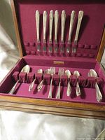 Full view of silver flatware set organized in wooden box with maroon fabric interior, showing knives in lid section and forks/spoons in bottom section.
