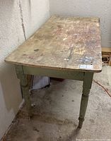 View of the antique pine harvest table showing the worn unpainted wood top, light green painted legs, and surface scratches with paint splatters.