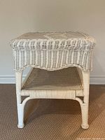 Side angle photo showing the antique white wicker end table with one lower shelf, woven wicker detail and leg construction.