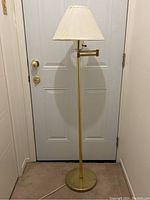 Full view of brass colored floor lamp with round base and fabric shade in an indoor setting, placed in front of a door.