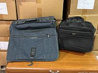Front view of navy blue Samsonite garment bag with multiple zippers and front pocket, alongside a black Delsey carry-on bag with dual handles showing.
