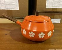Orange enamel fondue pot with teak handle and lid knob, white floral design around the pot body. Side view showing overall shape and design details.