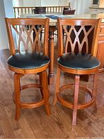 Pair of wooden counter height barstools featuring decorative wooden back designs and black faux leather padded seats with brass nailheads.