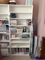 Front view of the white melamine bookcase filled with various household items (not included). The bookcase appears structurally sound but shows wear on the bottom shelf and Velcro strips on the top.