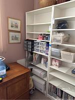 View showing white melamine bookcase in corner of room with contents on shelves including clear plastic drawers, baskets, and craft supplies. Some scuff wear visible on bottom shelf.