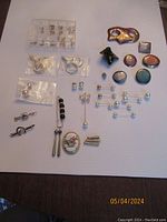 Top-down photo showing belt buckle, colorful pins, stud earrings, barbell pins, and bolo tie piece all arranged on white background.