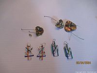 Full lot of five earrings shown including two pairs of geometric wire earrings in blue, green, orange, and gold, and three flower-shaped wire earrings in metal, one small and two larger with patina. One earring missing stud.
