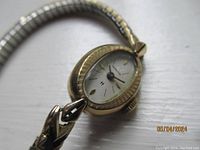 Close-up photo of the watch face showing oval gold-tone bezel, textured edges, cream dial, and gold markers and hands.