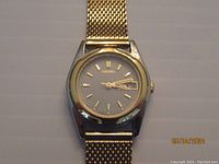 Front view of Seiko ladies watch showing grey dial, gold-tone hour markers, date window, and metal mesh band.