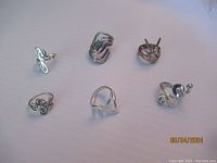 Photo showing six sterling silver rings with various designs; includes a hand-shaped ring, a heart-shaped ring with swirls, and rings with abstract looping designs.