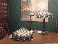Pair of lamps on wooden table, one lamp missing shade, showing bronze-tone base and columnar stand; Tiffany-style stained glass shade with colorful geometric floral design.