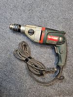 Image depicts green and black Metabo hammer drill with power cord coiled beside it on a grey surface. Label clearly shows Metabo brand and specifications.