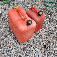 Two plastic jerry cans on gravel surface, one orange and one red, both with black screw caps and yellow vent caps, viewed from above.