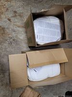 Two open cardboard boxes showing white disposable coveralls tightly packed and sealed in plastic with instruction sheets on top inside boxes.