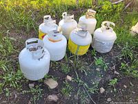 Seven 20lb propane tanks placed on the ground outside, some with wear and rust, untested operation.