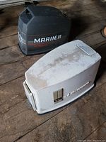 Photo showing black Mariner 25 HP boat engine cowl and white Johnson boat engine cowl on wooden floor, both with visible signs of wear and dust.