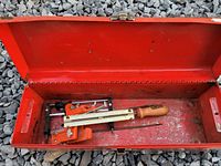 Open red metal toolbox containing chain saw sharpening tools including clamp-style filing guide and sharpening file.