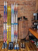 Four pairs of cross country skis in various brands and colors with five pairs of ski poles leaning against a wall beside a desk.