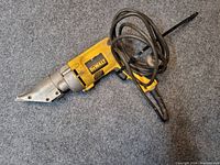 Angular view of the DeWalt electric metal shear tool lying on carpet, showing the blade and yellow body with attached power cord coiled.