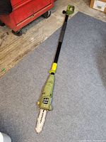 Full length view of the extendable electric pole saw, green with black and yellow parts, placed on a floor mat.