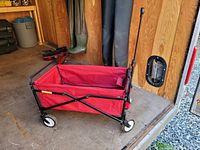 Red fabric folding utility wagon standing open with four white and black wheels, black metal frame, and telescoping black handle.