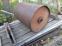 Photo showing a large 3ft wide cylindrical metal lawn roller drum with rust on the surface, mounted on a gray metal frame, photographed outdoors on a wooden surface near trees.