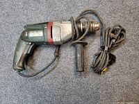 Full view of Metabo electric drill with attached side handle and coiled power cord lying next to it on a gray carpet surface.