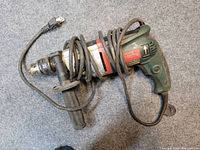 Top view of green and silver Metabo 750 hammer drill with attached side handle and coiled power cord on a grey carpeted surface.