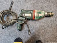 Photograph showing a Metabo electric hammer drill with green and red housing, cord attached and plug, showing signs of heavy use.