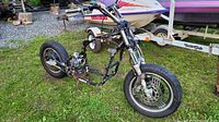 Full view of salvage motorcycle showing black frame, front forks, front and rear wheels, partial engine and exposed mechanical parts, placed on grass outdoors alongside boat trailer.