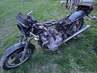 View of complete salvage motorcycle frame resting on grass, showing front fork, engine, seat frame, and rear section with wiring and lights.