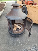 Outdoor metal fireplace with top vented chimney, mesh door open showing three small pieces of wood inside. Surface exhibits rust and wear.