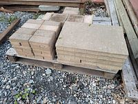 Full view of two pallets stacked with smaller square pavers on the left and larger rectangular slabs on the right, resting on gravel surface.