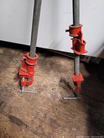 Two pipe clamps standing upright showing long metal rods with red clamp heads and T-handle screws for tightening
