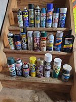 Photo showing three rows of various aerosol cans and one plastic bottle on wooden stairs including spray paints, lubricants, bug sprays and car wax products