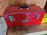 Closed red metal Stanley toolbox with black handle and two metal latches on a wood surface.
