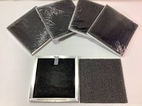 Five sealed square replacement filters arranged side by side with one filter unwrapped showing black filter material inside a white frame, plus a grey mesh sheet.
