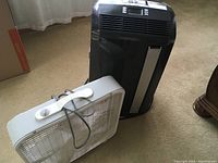 Two items on carpeted floor: black portable DeLonghi AC and white plastic box fan