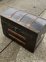 Photo of the vintage black dome top chest showing front and side details including metal hinges, wooden slats, and metal handles.