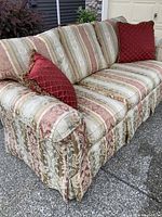 Front angle showing striped gold, red, and beige brocade upholstery and two red decorative pillows with fringe trim.