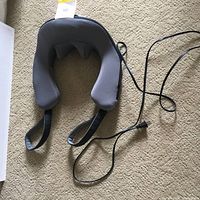 Top view of grey fabric neck massager with attached power cord laid on tan carpet.