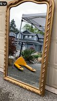 Full view of the large rectangular gold framed mirror leaning against a garage door outside showing overall shape, size, and scalloped top edge.