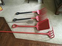 Five red and black food grade plastic and metal processing tools laid on carpeted and hardwood floors, including scoop shovel, pruning forks and paddle